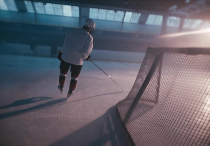 Hockey player circling a goal with a hockey stick on an indoor ice rink with a rising sun streaming through windows in the background