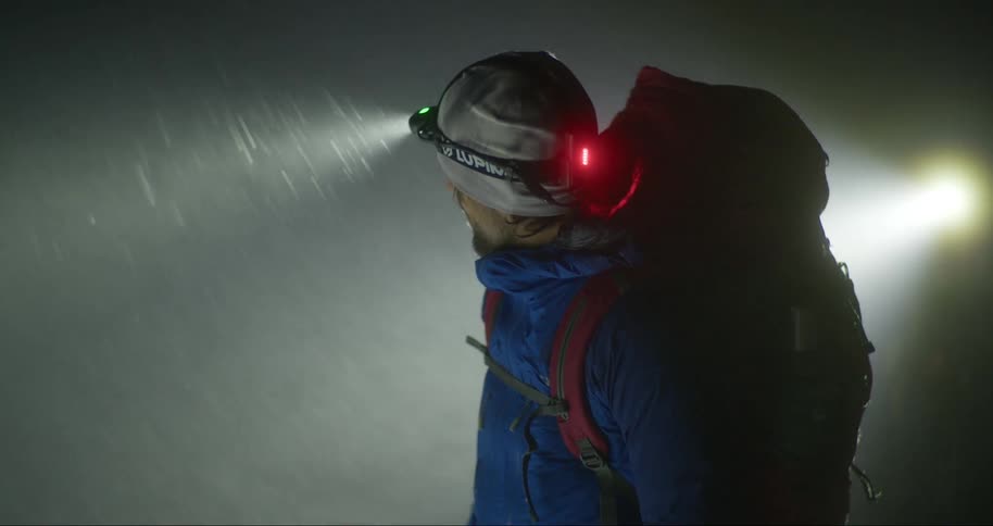 A man standing in the middle of a snowstorm on a mountain using a led headlamp