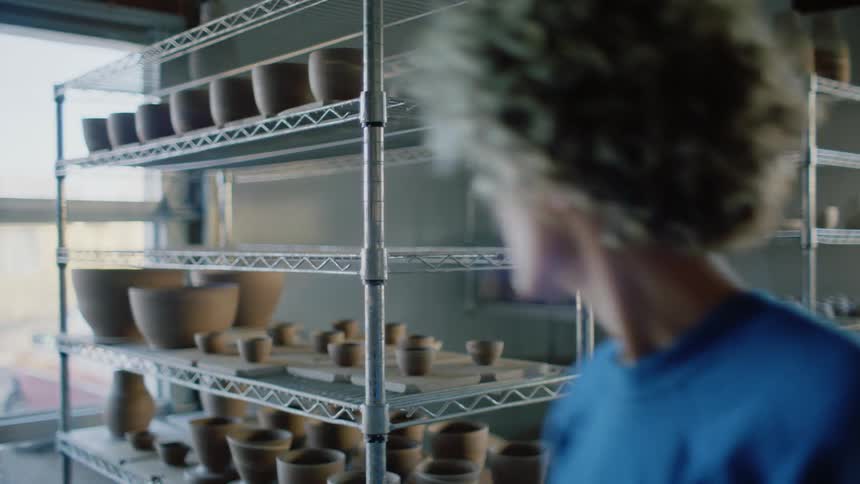 Female potter moves clay pots on squares from metal shelving unit