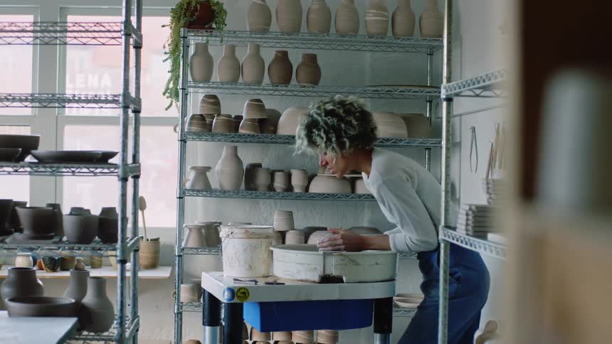 A standing woman is modeling clay and there is a shelf behind her full of vases