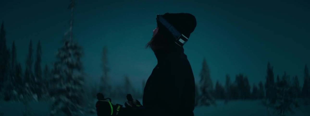 A woman in ski gear and a flashlight on her head stands on a snowy field and looks around looking scared