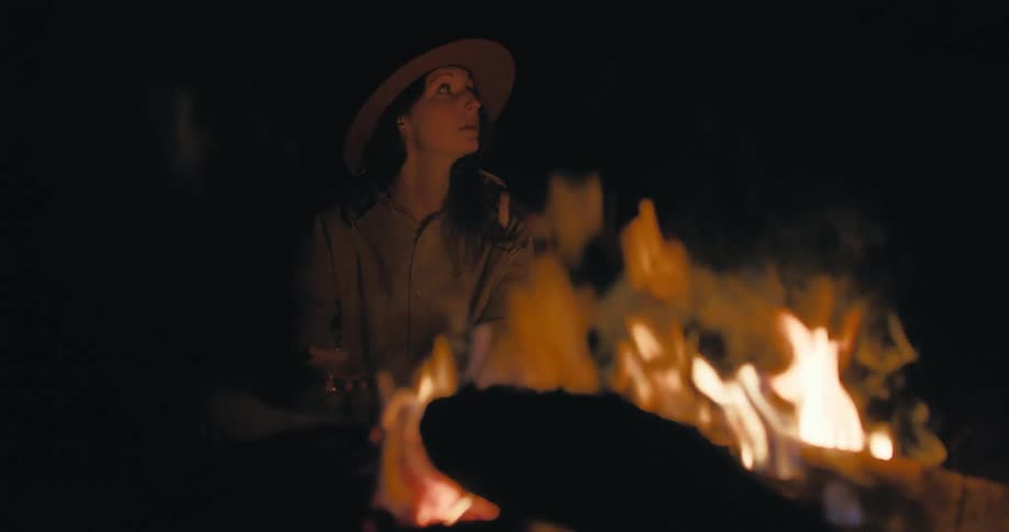 A woman sits outdoors at night next to a burning campfire