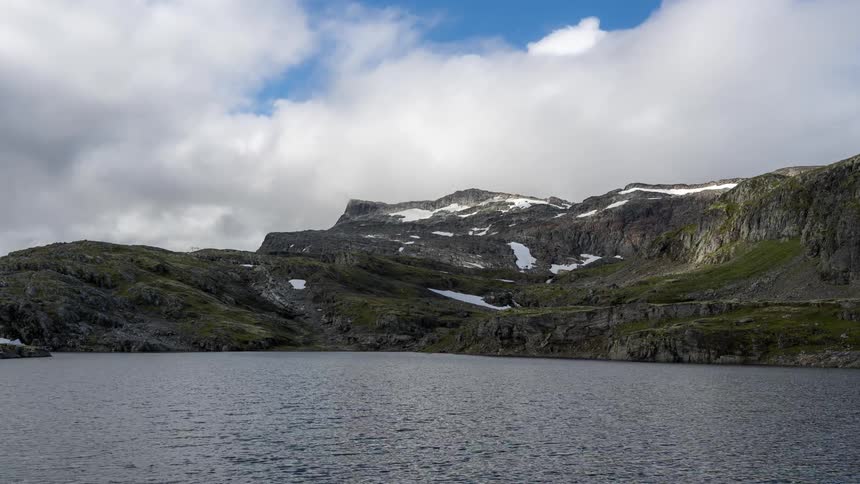 A landscape of a lake facing a mountain with some snow on top and many clouds in the sky