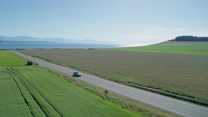A car drives between green crop fields with ocean and mountains in the distance