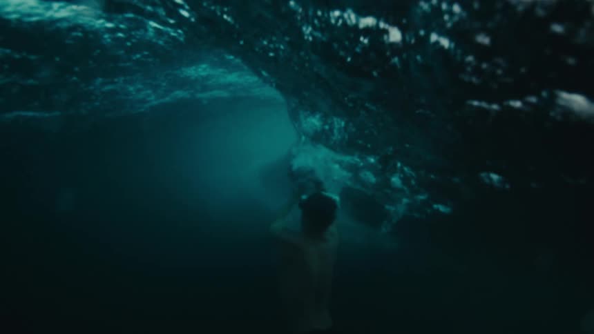 A man with a camera in his hand looking at a wave that is breaking and then he dives and seen underwater with some fish