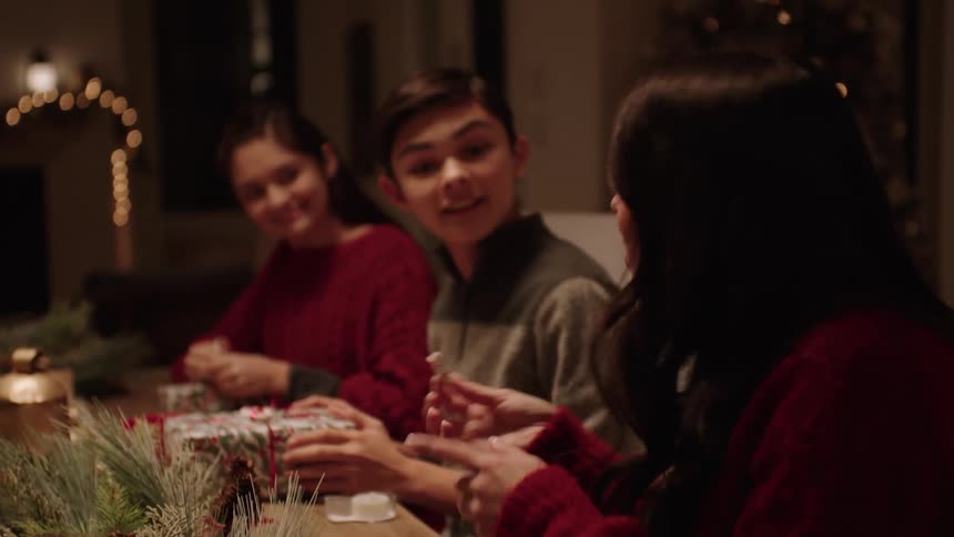 Family members sitting at tables with some eating and others assembling gifts during the festive season
