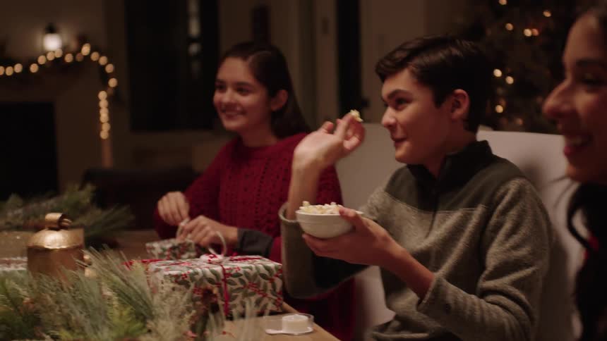 People seated at the dining table at night with a boy throwing popcorns in the mouth of a man while they all laugh