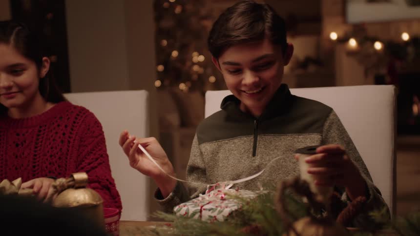 Male teen seated at the dining table talking and laughing while wrapping gift at night