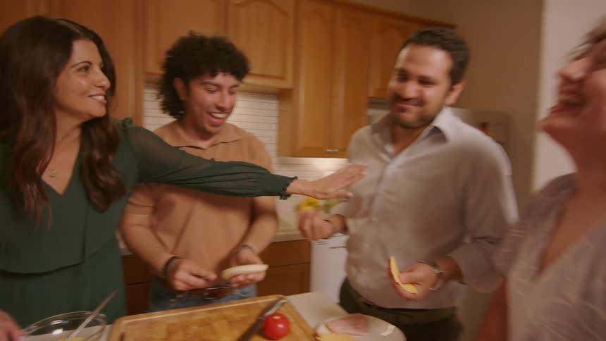 Four people are in the kitchen talking and smiling and a man takes a piece of cheese and eats it and everyone laughs