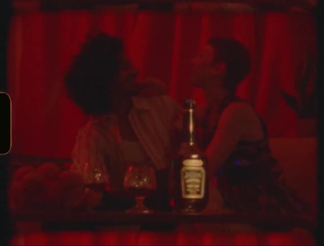 Woman leans against man at table holding bottle of liquor and goblets
