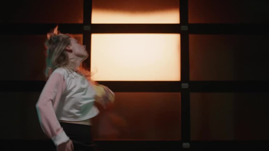 A woman dancing frenetically in front of a wall panel divided in rectangles backlit by orange light