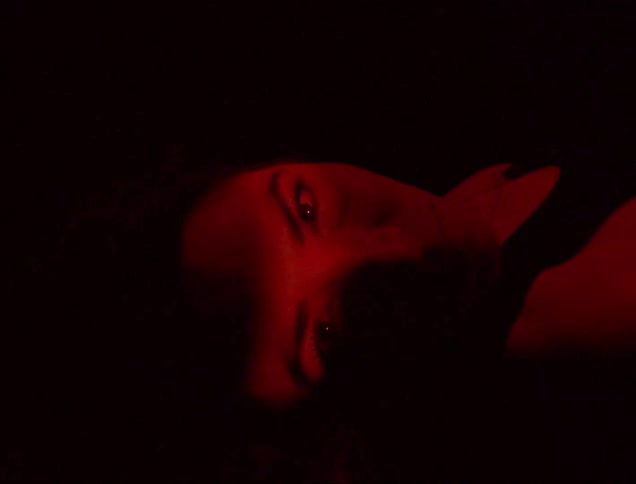 A woman lying down has her eyes closed and when a light hits her eyes she opens them and everything is red around her