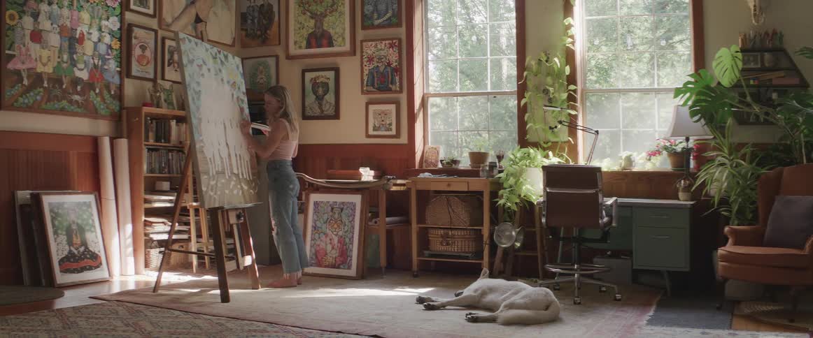 Female artist paints large easel while her dog sleeps on floor nearby