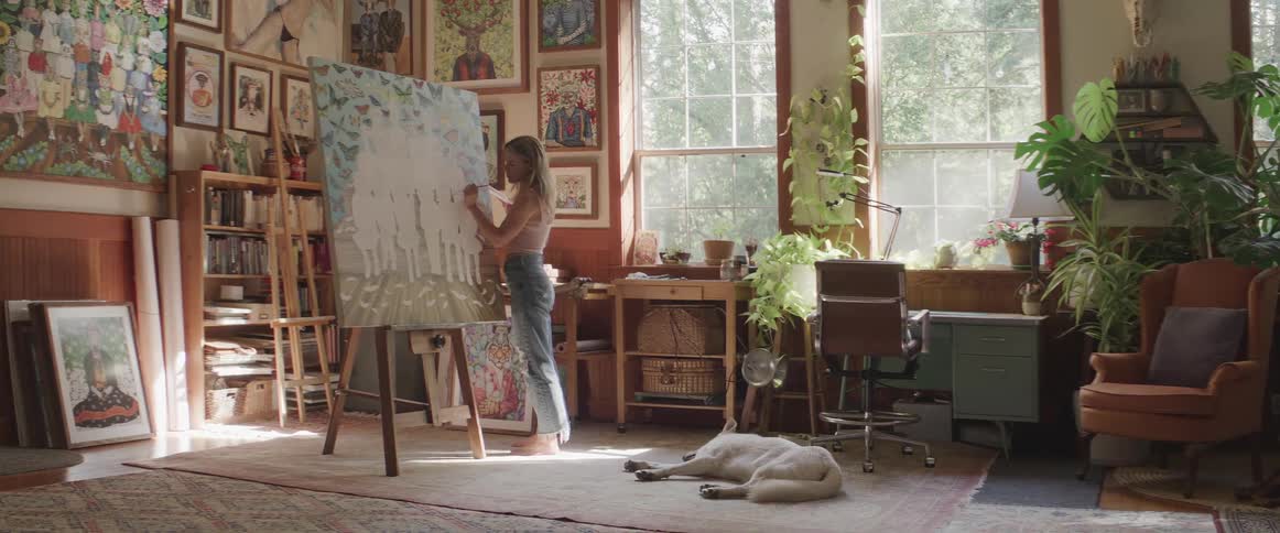 Blonde female paint large canvas indoors near window as dog lays on floor