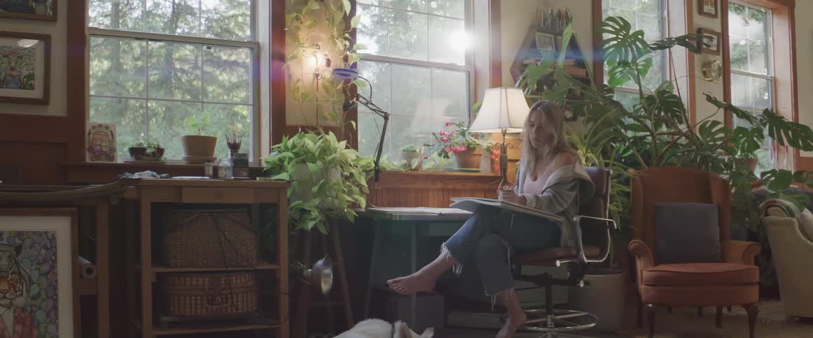 Blonde woman sits in room with multiple plants and draws in large sketchpad