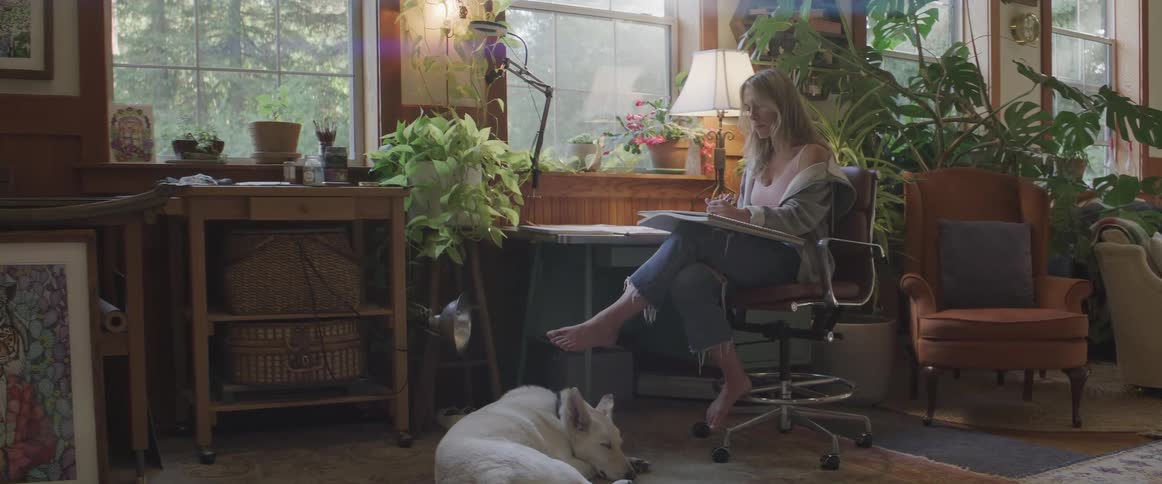 Blonde woman sits at desk indoors sketching on pad dog lays on floor