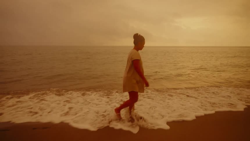 A young Asian woman explores a beach barefoot as waves roll in under a yellow sky