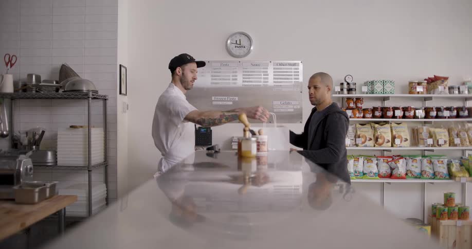 A man inside the counter hands a bag to a man who bought something from the establishment