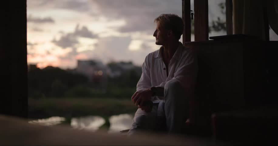 A man operating his mobile phone while seated by the window indoor at sunset