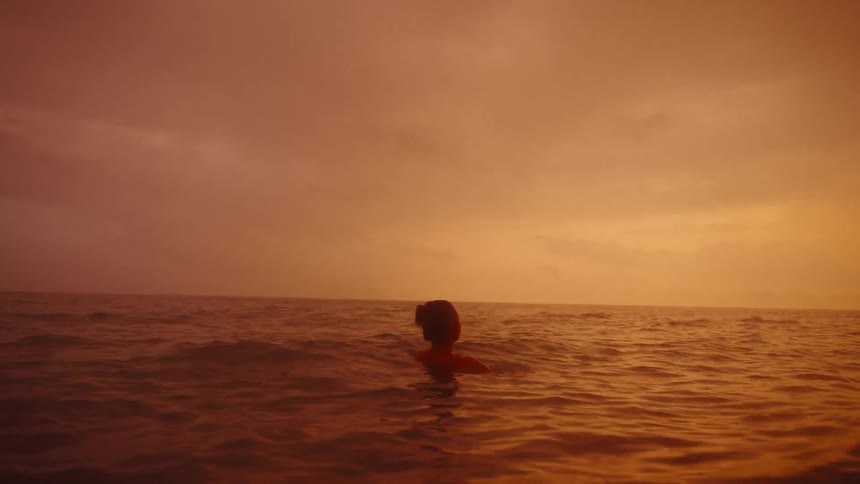 A young Asian woman floats in the ocean in the late afternoon under a dramatic red sky