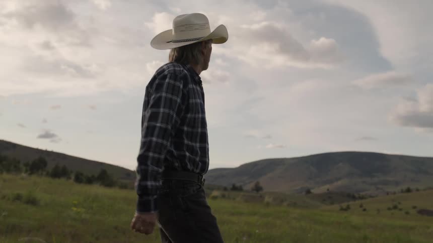 Elderly man wearing a cowboy hat walking on a hill next to mountains during the day