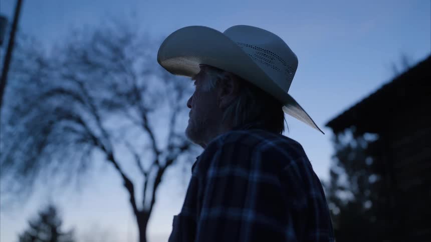 An old man wearing a cowboy hat outside of his home