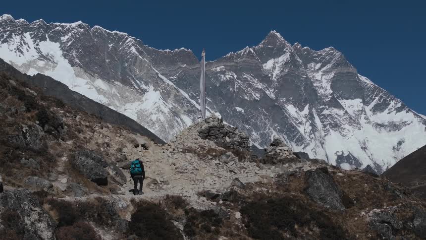 A person with a large backpack is walking on a mountain towards a flag that is moving due to the wind and behind is a huge mountain with snow on top