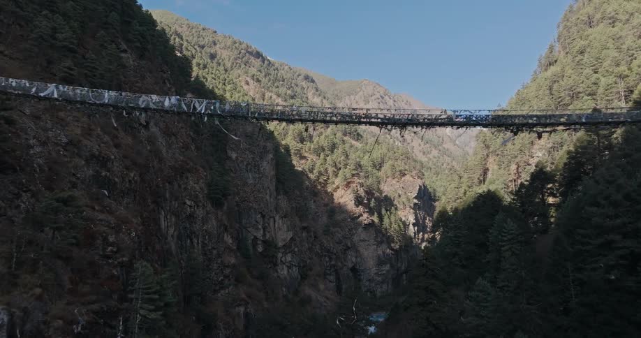 Person carrying package crosses one of two footbridges over river in valley toward side with blue roofed building