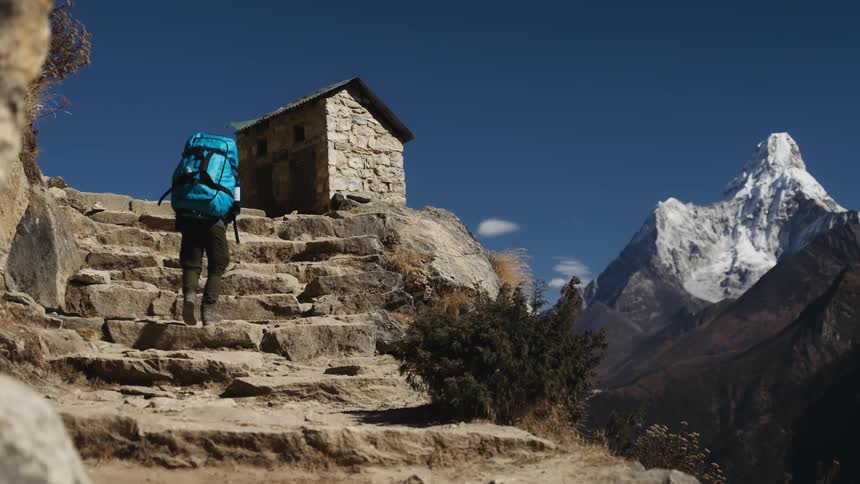 A man with a large backpack is climbing stone steps to a small hut and back to a huge mountain with snow on top