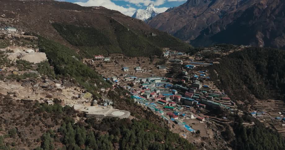 Blue and red roofed town buildings occupy mountain slopes near conifer forest on a cloudy day