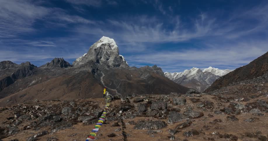 A hiker walking along a rocky plateau and passing a wire with flags hanging from it overlooking a snow mountain range