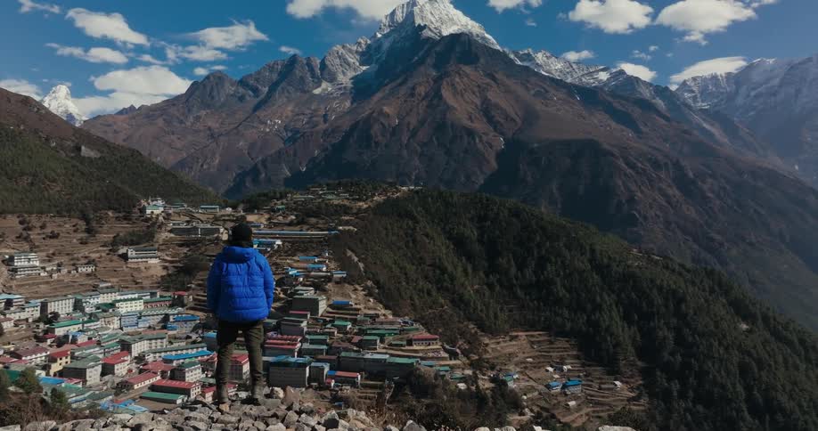 A person in a blue jacket has his back turned and observes a city built at the base of a mountain
