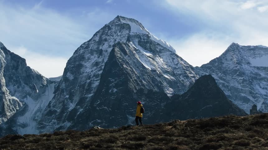 A hiker walks along a ridge line in front of a steep mountain range on a partly cloudy day