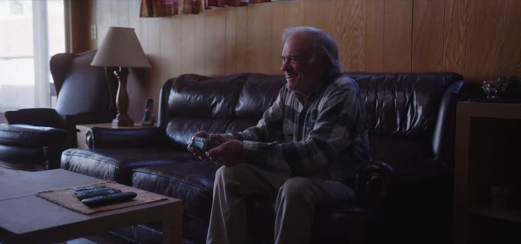 Elderly man with a plaid shirt and brown pants sits on black couch playing video game