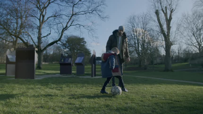 A father watches his young son kick a soccer ball around a park