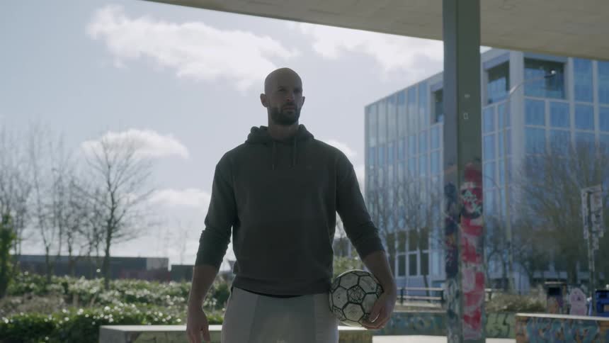 Bald man stands under a pavilion holding a soccer ball on a cloudy day