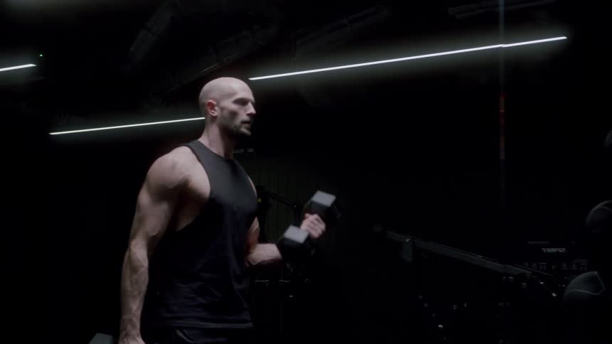 Bald man lifts weights while standing in dark gym