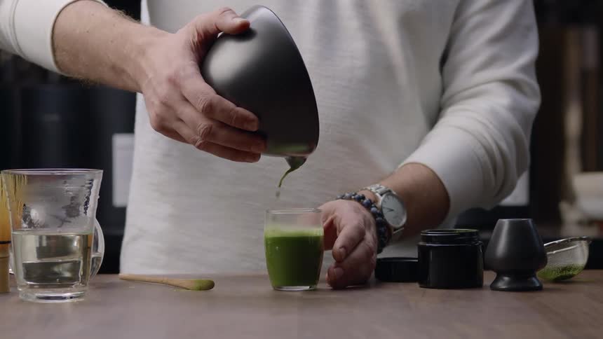 Hands of a man pouring from a small size a liquid of a green juice into a glass cup