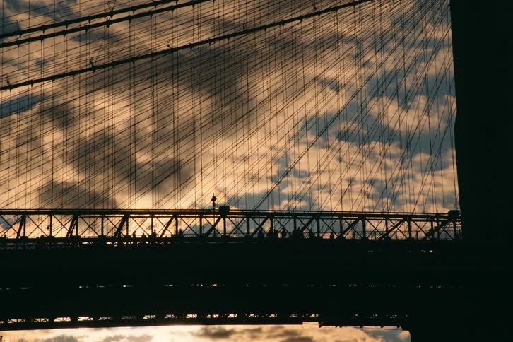 People crossing suspension bridge are silhouetted against the cloudy sky at sunset