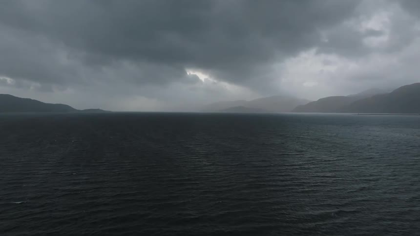 Waves flow across water away from storm clouds and fog on horizon between hills on shore