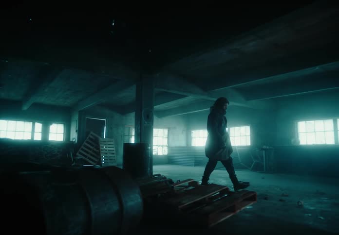 A solitary person walks through a room in an derelict building and stands at a fire