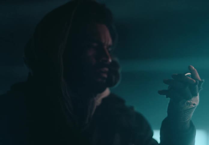 A hooded man inspects a ring inside a dimly lit room