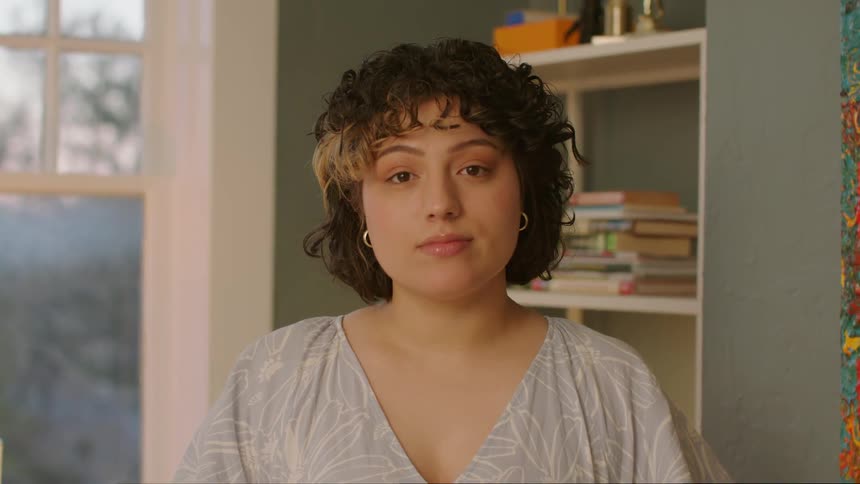 A woman standing in a room next to a window turns to look at the camera