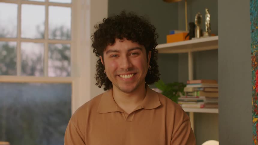 A man with curly hair a nose piercing earrings and a brown polo shirt smiles at the camera
