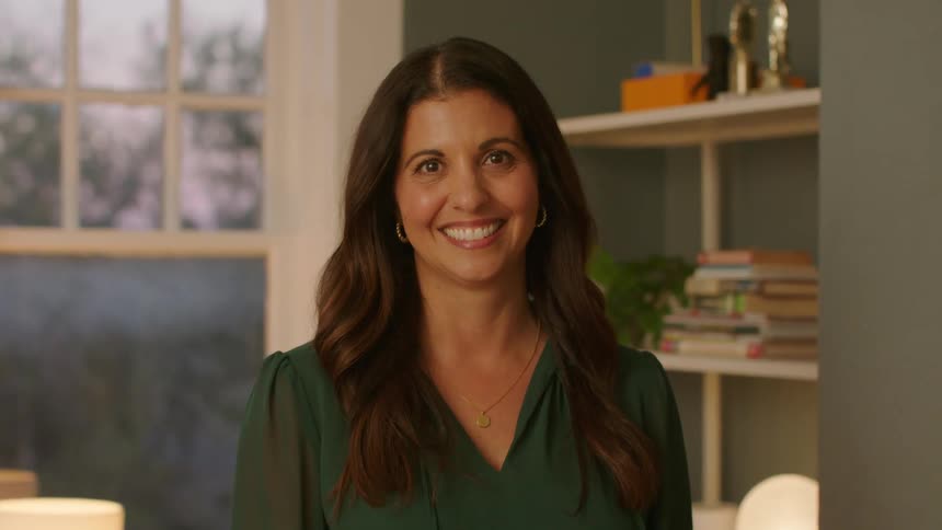 Woman with brown wavy hair and a green shirt smiles at camera in a room