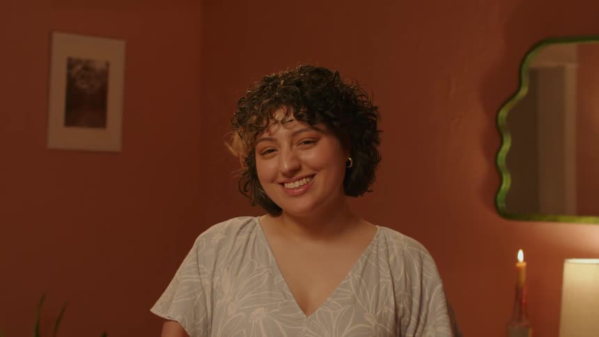 Woman with curly hair earrings and light blue blouse smiles at camera in a room with orange walls
