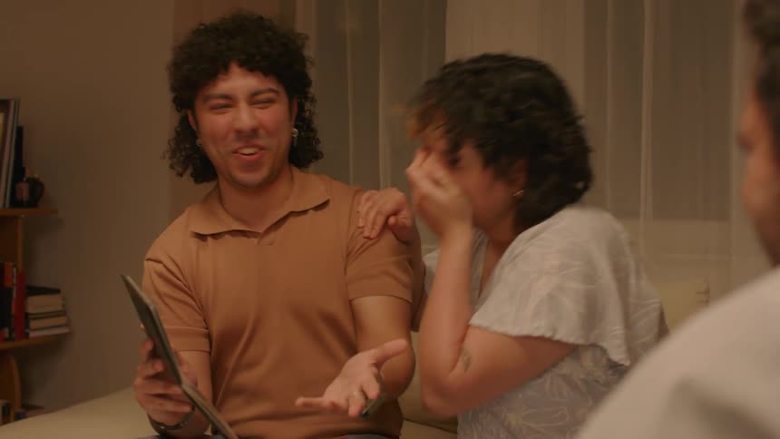 A couple are sitting and showing a photo to a person in front of them and that person points to the photo and everyone laughs