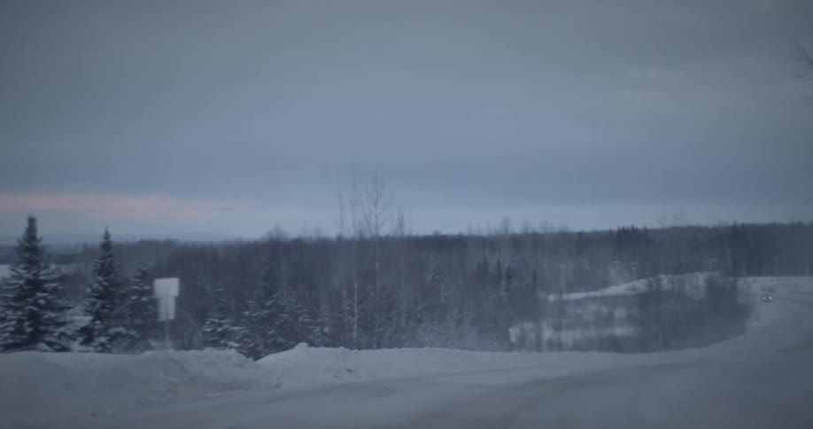 Panorama of a snowcovered road with a car passing through surrounded by coniferous trees in a cloudy morning