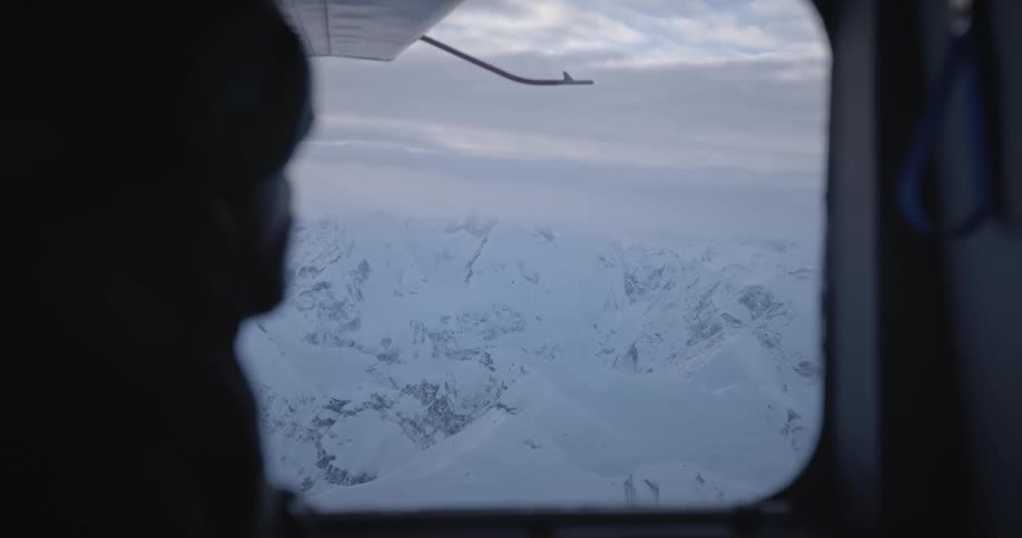 A man is sitting and looking out of the window of an airplane flying over snowy mountains