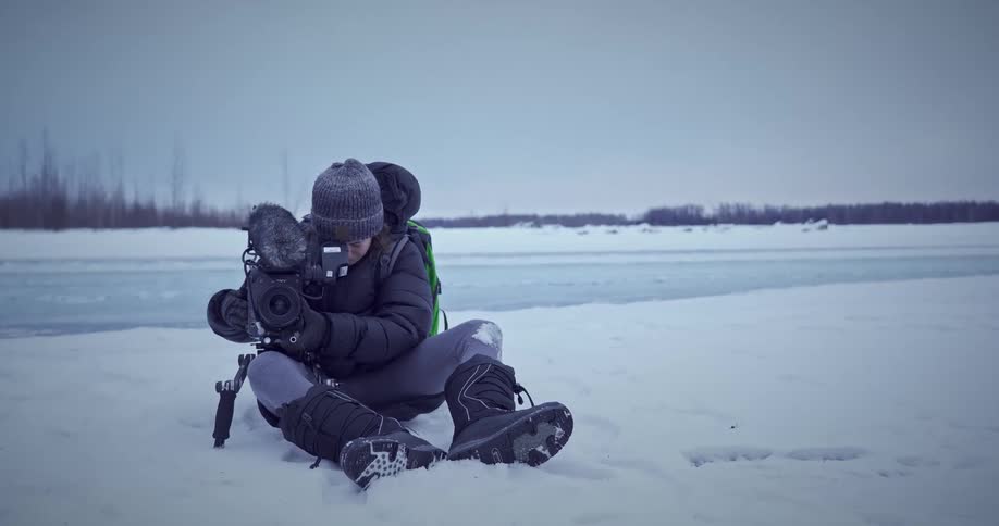 A person sitting in the snow by a frozen lake adjusting a camera
